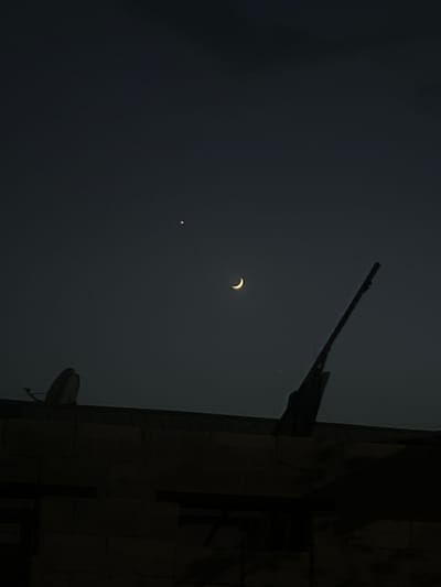 This image captures a celestial event, likely a partial solar eclipse, viewed from Balkh, Tajikistan, at dusk or dawn. The prominent subject in the sky is a crescent-shaped object, illuminated from the right, suggesting a portion of the sun is obscured. To the lower left of the crescent, a small, bright point of light is visible, which could be a planet or a star.
The foreground is dominated by a dark, silhouetted structure on the right side of the frame. This appears to be a building with architectural details, possibly stone or brick, exhibiting a textured surface and projecting elements that resemble gargoyles or decorative protrusions. The dark sky suggests low light conditions, reinforcing the idea of twilight. The overall atmosphere is somber and mysterious, emphasizing the celestial spectacle. No text is visible in the image.