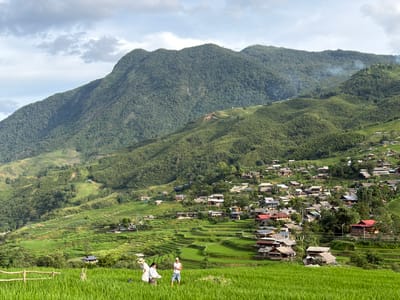 A wide shot captures the verdant beauty of Tả Van village in Vietnam, nestled amongst rolling hills and terraced rice paddies. The lush green fields dominate the foreground, leading the eye to a cluster of houses that form the village, built on the slopes of the mountains. The houses, a mix of traditional and modern architecture, are nestled among trees, with red-tiled roofs a common feature. Smoke rises from a few chimneys, indicating daily life is in full swing. In the mid-ground, two individuals are engaged in activity within the rice field, their presence adding a human element to the sweeping landscape. The background is defined by the imposing presence of the mountains, covered in dense greenery, under a sky filled with scattered clouds. The overall impression is one of tranquility and harmony between human settlement and the natural environment.