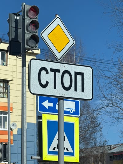 The image shows a set of traffic lights and several road signs in Beloyarsky, Russia. The traffic lights display a red light. The road signs include a priority road sign (yellow diamond), a "СТОП" (STOP) sign in Cyrillic, a bus stop sign, and a give way sign. Bare tree branches and a bright blue sky are visible in the background. The structures of buildings are also present in the background.