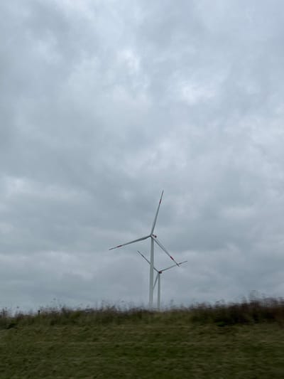 The image displays a landscape in Dornstadt, Germany, under an overcast, grey sky. Two wind turbines are the primary subjects. The foremost turbine, centrally located towards the right, consists of a tall white tower with three white blades, each tipped in red, oriented approximately at 11 o'clock, 3 o'clock, and 7 o'clock positions. A second, partially obscured wind turbine is situated behind and to the right of the first, exhibiting similar construction. The foreground features a blurred expanse of green and brown grassy terrain, indicative of motion by the camera's perspective. No individuals or discernible activities are present. The environmental lighting is diffuse and consistent with dense cloud cover.