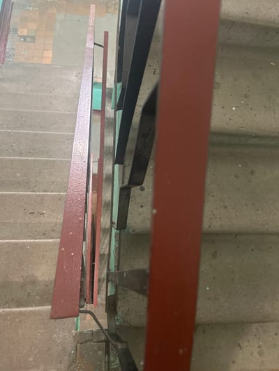 This image depicts a set of concrete stairs with a red metal railing on the left and a dark metal railing with green accents on the right. The stairs appear to be outdoors or in an industrial-style building, with some dirt and debris visible on the steps. The perspective is looking down the stairs. The location is identified as Posyolok Imeni Morozova, Russia. There are no people or any discernible actions taking place in the image.