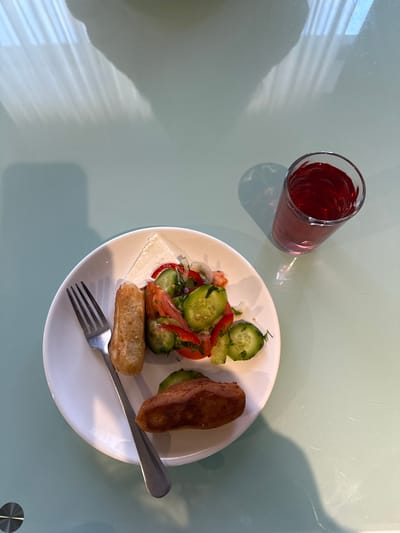 The image displays a meal set on a light green tabletop in Bakuriani, Georgia. A white circular plate, positioned in the lower left quadrant, holds various food items. On the plate, a triangular slice of white cheese is visible, alongside a fresh salad composed of sliced cucumbers, tomatoes, red bell pepper strips, and chopped green herbs. Two fried food items are also present: one is a cylindrical shape, and the other is an oblong, flatter shape. A silver-colored four-tined fork rests on the left side of the plate. To the upper right of the plate, a clear glass contains a reddish-purple liquid. The tabletop exhibits reflections of vertical window blinds or slats originating from the upper left. Shadows of the plate, the glass, and an indistinct, larger overhead object or person are cast on the table surface. No individuals are visible in the scene.