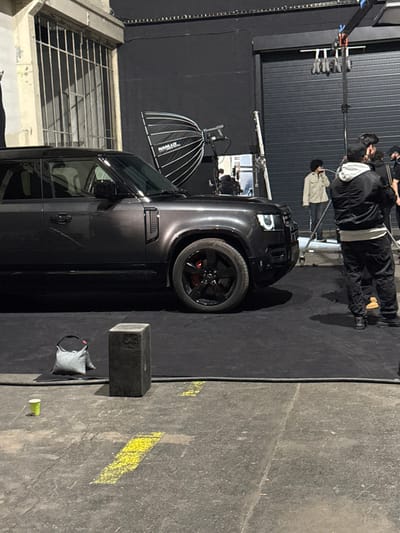 The image captures an indoor production shoot scene within an industrial or studio facility in Ivry-sur-Seine, France. A dark grey Land Rover Defender SUV, positioned left-of-center, is the primary subject, resting on black matting. The vehicle's front passenger side is visible, featuring black multi-spoke wheels with red brake calipers and illuminated rectangular daytime running lights.

To the right of the SUV, a male individual stands with his back to the camera, wearing a black bomber jacket with a grey hooded sweatshirt underneath, dark pants, and dark shoes. Further to the right and slightly behind him, a second male individual, dressed in a light-colored jacket and dark pants, holds a microphone and faces right. Additional figures and equipment are partially visible in the background, including another individual seated in what appears to be a director's chair behind the SUV.

Behind the vehicle, a large Nanlux parabolic softbox on a stand is positioned as a light source, casting reflections on the vehicle's surface. Overhead rigging with multiple small lights is visible. A large black roll-up garage door dominates the background behind the main subjects. To the far left, a multi-paned window with metal bars is visible, set into an aged, light-colored wall.

The foreground features a concrete floor with worn yellow painted lines, partially covered by the black matting. A dark, rectangular block and a grey weight bag are positioned on the matting. A light green disposable cup rests directly on the concrete floor to the left of these objects. The environment indicates a controlled setting for filming or photography, utilizing professional lighting and staging elements.