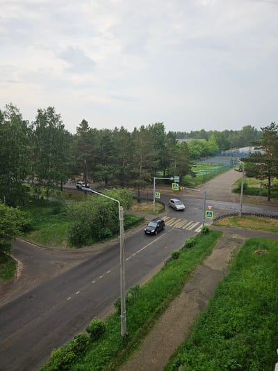 The image shows an aerial view of an intersection in Sosnovoborsk, Russia. The sky is overcast. On the road, a silver car and a black car are visible. A pedestrian crossing is marked on the road. Traffic lights and street signs are present. Trees and grassy areas surround the intersection. In the background, there is a playground and a fence.