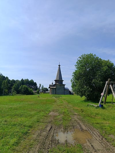 The image depicts a rural scene in Semenkovo, Russia, featuring a traditional wooden church as the central subject. The church, constructed of dark wood, has a distinctive tiered roof topped with a spire. In front of the church, a muddy track with tire marks and a puddle cuts through a vibrant green grassy field dotted with small yellow wildflowers. To the right of the church and field, a large, lush green tree dominates the landscape. Adjacent to the tree, a wooden swing set with two upright posts and a horizontal bar is visible. In the background, a dense line of green trees forms a horizon, with glimpses of some low-lying buildings peeking through. The sky above is a clear blue with sparse, wispy white clouds. The overall environment suggests a sunny day in a tranquil, natural setting.