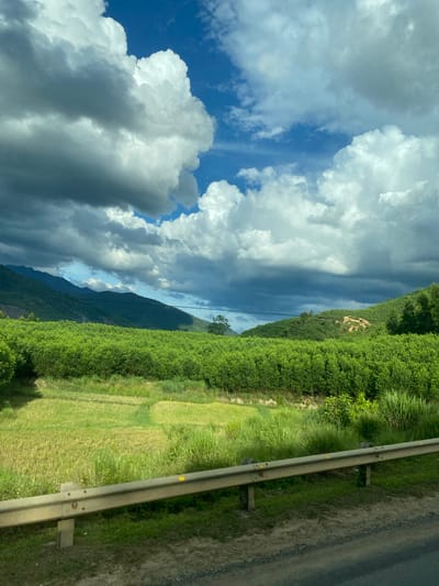 In the image, a vast green field stretches towards a backdrop of rolling, tree-covered hills under a dramatic sky filled with a mix of dark and white clouds. In the foreground, a metal guardrail runs along the edge of a road. The landscape suggests a rural setting, likely in Thôn 18, Vietnam, characterized by lush vegetation and natural topography. No people or specific actions are depicted, focusing instead on the environmental panorama.