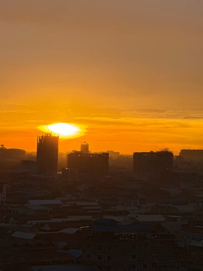 This is a photograph taken at sunset in Khan Mean Chey, Cambodia. The sun, a brilliant orange orb, is low on the horizon, casting a warm, hazy glow across the sky. Wisps of clouds are visible, tinged with the same golden hues.

In the foreground and stretching across the right side of the image is a city skyline dominated by silhouetted buildings. These structures appear to be a mix of residential and commercial buildings, with some under construction, evidenced by scaffolding on one of the taller buildings. The architecture is modern, with sharp lines and flat roofs.

The overall mood of the image is tranquil and serene, capturing the peaceful transition from day to night. The setting is urban, suggesting a populated area within the city. The time of day is clearly sunset, given the intense orange light and the low position of the sun. There is no visible text in the image.