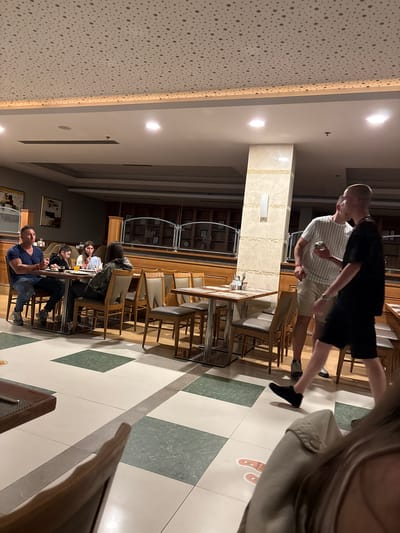 This media file captures an indoor scene, likely a hotel restaurant or dining hall in Selçuk, Turkey, during the evening or night. The space is illuminated by warm artificial lighting, including recessed spotlights and long strip lights along the ceiling.
The foreground and mid-ground show two men walking from right to left across a floor with a distinctive pattern of white and green/grey square tiles. One man, closer to the camera, wears a black t-shirt and black shorts, with black shoes. The man in front of him wears a white and grey striped t-shirt, light-colored shorts, and holds a small, light-colored object in his hand. Partially visible in the extreme foreground on the right, what appears to be the camera holder's shoulder and arm (wearing a light-colored jacket) is seen. On the floor near the foreground, a red and white decal, possibly a footprint, displays the partially visible text "KID'S BUFFET," suggesting a buffet area nearby.
In the background and to the left, several people are seated at tables, engaged in dining or conversation. One man, prominent in a dark blue t-shirt and jeans, sits with another person (possibly a child) in a light top, and a woman with dark hair in a patterned jacket. The tables are set with drinks and other items. The furniture includes modern chairs with light upholstery and wooden frames.
The architectural features include a large, polished marble-clad pillar in the center-right of the frame. The ceiling on the left features a white, perforated panel design, while the right side has smooth white panels with integrated lighting. Wooden paneling and shelves with various items are visible on the far-left wall, along with some framed artwork. The overall atmosphere appears casual and relaxed.