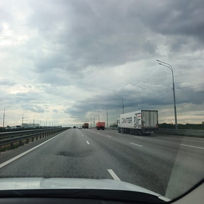 The image shows a cloudy sky over a highway. A white truck with "CRAFTER" written on its side is driving on the highway. Other vehicles and streetlights are also visible. The scene appears to be on a highway with multiple lanes and a guardrail. In the distance, buildings and trees can be seen. The camera is positioned from inside a vehicle, looking forward. The location is identified as Mikhnevo, Russia.