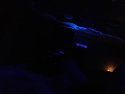 The image depicts a predominantly dark interior environment, illuminated primarily by blue and faint orange light sources. In the upper right quadrant, irregular, wave-like or cloud-like patterns of bright blue glow are visible on what appears to be an overhead surface or wall, suggesting illumination by blue or ultraviolet light. Below this, two distinct horizontal linear blue light sources are present in the mid-ground. Towards the lower right, a small, distant, and dim orange light source is discernible. No individuals are visible. The overall scene, characterized by dim lighting and blue thematic illumination, is consistent with an exhibit area designed to simulate an underwater or nocturnal environment. This setting strongly suggests an interior gallery within the **Aquarium of the Pacific in Long Beach, United States**, likely a deep-sea or bioluminescent exhibit.