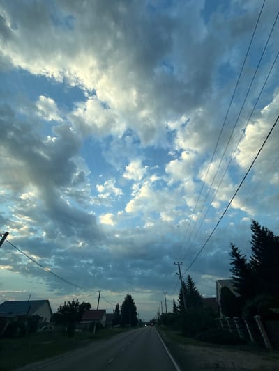 The image depicts a rural road lined with houses and trees, under a dramatic sky filled with scattered clouds. The perspective is from within a vehicle, looking forward and slightly upward. The road itself appears to be asphalt, with a white line marking the edge on the right side. Overhead, multiple power lines stretch across the frame, supported by wooden poles. On either side of the road, houses with visible roofs and some vegetation are present. The sky is a mix of blue patches and large, textured clouds, some illuminated by sunlight, giving the scene a dynamic, possibly late afternoon or early evening atmosphere. The presence of power lines and the style of housing suggest a residential area in a village or small town. The general impression is one of a quiet, everyday scene.