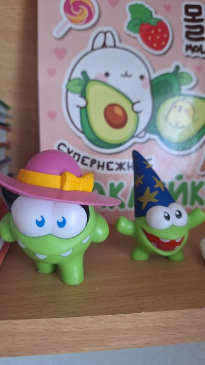 Two green cartoon characters are on a wooden shelf. The character on the left is wearing a pink hat with a yellow band and bow. The character on the right is wearing a blue wizard hat with yellow stars. The background is a pink surface with cartoon images of avocados, a strawberry, and a lollipop. The image is taken from a slightly high angle. The exact location in Nogliki, Russia cannot be determined from the provided image.