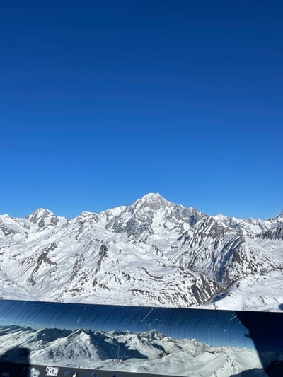 The media file presents a striking aerial or high-altitude view of a vast, snow-covered mountain range under a brilliant blue sky, likely in the French Alps near Séez. The image, if rotated 90 degrees counter-clockwise for correct orientation, shows the clear, intense blue sky dominating the upper half, with a subtle gradient from a deeper blue at the top to a slightly lighter hue towards the horizon.
The lower half of the image is filled with rugged, majestic mountains, heavily blanketed in pristine white snow. Exposed dark rock faces and dramatic peaks and ridges are visible, indicating deep valleys and a complex topography. The strong sunlight illuminates the snow, creating sharp contrasts and highlights that emphasize the textures and contours of the landscape.
In the right foreground, a dark, reflective surface, resembling a window or part of a vehicle interior (possibly an airplane or helicopter), is visible. This surface displays reflections of additional snow-covered terrain, extending the perceived depth of the mountainous scene. Within this reflective area, a small rectangular shape with faint white text "HP" is discernible, suggesting a laptop screen or a device label.
The scene depicts a bright, clear daytime with excellent visibility and sunny weather. There are no clouds in the sky, highlighting the vastness and serenity of the alpine environment. No people are visible, emphasizing the grandeur and untouched nature of the landscape. The overall impression is one of awe-inspiring natural beauty and immense scale.
