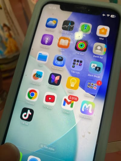 An iPhone, contained within a light blue/mint green protective case, is depicted in an indoor setting in Đak Đoa, Vietnam. A human finger is partially visible in the bottom left foreground, contacting the lower edge of the phone's screen. The iPhone display is active, showing its home screen with multiple application icons arranged in a grid. The top status bar indicates a time of 14:45, airplane mode is active, Wi-Fi is connected, and the battery level is 66%.
Visible application icons include: Thời tiết (Weather), Chứng khoán (Stocks), Podcast, Sách (Books), Thể dục (Fitness), Nhà (Home), Tệp (Files), Xem trước (Preview), Danh bạ (Contacts), Freeform, Nhật ký (Journal), Tiện ích (Utilities), Dịch thuật (Translate), Chrome, YT Kids, Meo (Tips), Gmail (displaying 3,916 unread notifications), Giáo dục (Education, a folder containing apps, showing 1 notification), TikTok, and MiraiMind. A search bar labeled "Q Tìm kiếm" (Search) is present at the bottom of the screen. The screen surface shows minor smudges and dust. In the blurred background, a printed image or book cover featuring a female character in a yellow gown is partially visible.