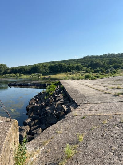 The image depicts a sun-drenched scene along a waterway, likely the Dniester River at its confluence with the Prut, near Ustia, Moldova. The day is clear and bright, suggesting mid-morning or early afternoon.
The foreground is dominated by a large, weathered concrete structure, possibly part of a dam or industrial embankment. This structure angles sharply from the right side of the frame towards the center, showing its rough, granular surface with patches of dry grass growing through cracks. Below and to the left of this concrete platform, a jumble of dark, angular concrete blocks forms a breakwater or reinforcement for the waterway.
The waterway itself is a calm, murky expanse, reflecting the blue sky and the lush green foliage on the opposite bank. Algae or aquatic plants are visible on the surface of the water. Along the distant bank, a dense forest covers rolling hills, creating a vibrant green backdrop that stretches across the horizon. The trees appear healthy and full, indicative of a warm, growing season.
In the water, a long, dark structure, possibly a series of barges or a floating pier, is visible. The scene is devoid of any people, adding to a sense of quietude and perhaps abandonment. The overall impression is one of a functional, perhaps industrial, area integrated with a natural landscape. The clear sky and bright sunlight convey a feeling of a warm, summer day.