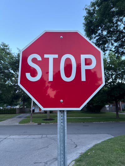 An octagonal red stop sign with white lettering is centered in the frame, mounted on a metal pole. The sign displays the word "STOP" in large, reflective white capital letters. The pole is visible extending downwards and out of the frame. Behind the stop sign, a suburban street is visible with a sidewalk and green lawns on either side. Trees with dense green foliage line the street, and a clear blue sky is present above. The scene appears to be set in the daytime, with the lighting suggesting it might be late afternoon or early evening. The overall environment is residential and peaceful. This stop sign is located at the intersection of Belvoir Boulevard and Edgewood Road in Shaker Heights, Ohio, United States of America.