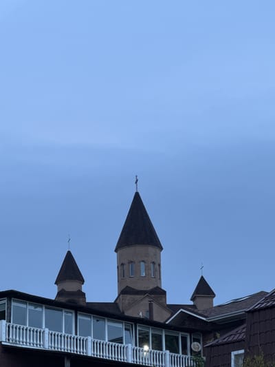 The image shows a building with a tower and steeple against a blue sky. The building has a dark-colored roof and beige walls with vertical windows on the tower. Adjacent to the tower is a section of the building with a white, decorative railing and multiple windows. There is also a smaller steeple to the left of the main tower. The photograph is taken from a low angle, emphasizing the height of the structure. The scene appears to be in Novomikhaylovsky, Russia, due to the architecture style, although this cannot be fully confirmed with certainty without further information.