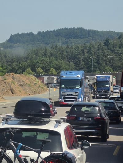 The image depicts a traffic jam on a highway near Katzenfurt, Germany. In the foreground, the rear of a white car with a bicycle rack and a black cargo box is visible. Next to it, a black Mercedes-Benz SUV with a license plate reading "LDK AO 9" is also in traffic. Further ahead, a blue DAF truck with "ANHALT LOGISTICS" branding is at the front of a line of vehicles. Behind the DAF truck, a blue Mercedes-Benz truck is also present. The highway is bordered by a steep, rocky embankment on the left and a dense forest of green trees on the right. The sky is clear and a pale blue. The scene suggests a typical highway traffic situation on a sunny day.