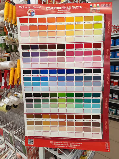 The image shows a close-up view of a color chart poster for "Kolerovochnaya Pasta" (Coloring Paste) from the brand Palizh. The poster is displayed in a retail setting, likely a hardware or paint store, given the presence of paint rollers and shelves with cans of paint in the background. The color chart features rows of paint swatches, each labeled with a number and a color name in Russian, such as "алый" (scarlet), "коралл" (coral), "персик" (peach), "оранжевый" (orange), "желтый" (yellow), "черника" (blueberry), "сиреневый" (lilac), "лаванда" (lavender), "слива" (plum), "аквамарин" (aquamarine), "бирюза" (turquoise), "синий" (blue), "фиалка" (violet), "ирис" (iris), "черный" (black), "хаки" (khaki), "лайм" (lime), "зеленый" (green), "изумрудный" (emerald), "бежевый" (beige), "кофейный" (coffee), "карамель" (caramel), and "шоколад" (chocolate). Some labels also include application instructions like "выкрасы на ЛАКЕ" (samples on varnish) and "выкрасы на КРАСКЕ" (samples on paint), along with percentages and ratios. At the bottom of the poster, there is text with company information, including the name "ООО «НОВЫЙ ДОМ»," address, phone number, and website: "WWW.PALIZH.RU, VK.COM/IPALIZH." A QR code is also visible. The overall impression is one of a commercial display in a store, showcasing a range of paint colors.