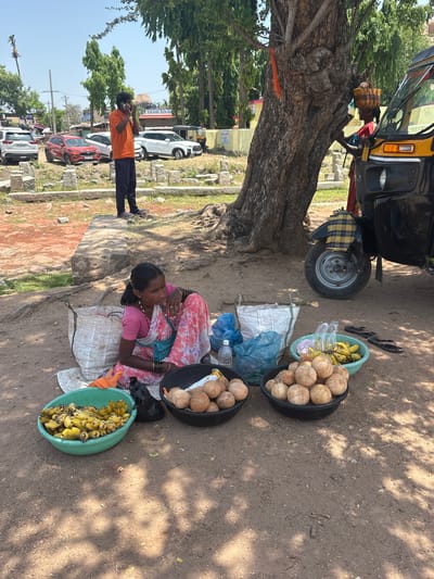 The image depicts a street scene in Kamalapura, India. A woman is sitting on a sack on the ground, surrounded by three large bowls containing coconuts and bananas. These items are likely for sale. Beside her is a parked black auto-rickshaw. In the background, a man in an orange shirt is standing on a street with parked cars. A large tree provides shade over the woman and her merchandise. The ground is dirt and scattered with leaves.