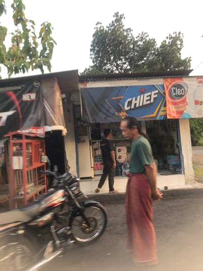 In Sentul, Indonesia, a blurry image captures a street-level view of a small storefront. A man in a green t-shirt and a red sarong stands in the foreground, his hands clasped. Behind him, another man in a black t-shirt and dark pants walks away from the camera. The storefront, adorned with a "CHIEF" banner and a "Cleo" advertisement, displays various products on shelves visible through the open entrance. A red cage-like structure with shelves stands to the left of the storefront, and a black motorcycle is parked in front of it, its details blurred by motion. Trees and foliage are visible in the background, against a bright, possibly overcast sky.