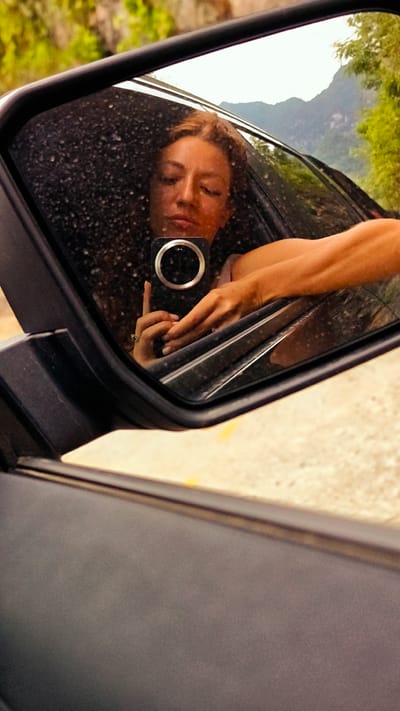 In Cán Tỷ Commune, Vietnam, a side mirror of a vehicle reflects a woman taking a picture of herself with a smartphone. The car appears to be on a road, with trees and hills visible in the background. The mirror shows raindrops on the glass surface. The woman's arm is resting on the window frame.