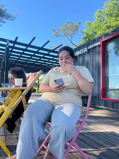 In Ochamchire, Abkhazia, a woman with dark hair and fair skin is seated on a pink chair, looking down at her phone and touching her mouth. She is wearing a light beige t-shirt and light blue pants. To her left, a man with dark, wavy hair is seated in a yellow chair, leaning forward and looking down. Between them on a yellow table is a white cup with a red design and a small pink object. In the background, a dark metal shipping container with "AMTA COFFEE" written on it in white letters is visible. A red-framed window is on the side of the container. The scene is outdoors, with a wooden deck and trees under a clear blue sky.