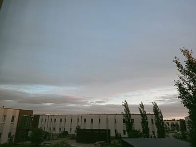 The image depicts an overcast sky above a low-rise residential building complex in Peetri alevik, Estonia. The sky is a blend of pale blue and grey with hints of pink and orange from what appears to be a sunset or sunrise. The buildings are modern, with white facades and dark-framed windows. Several trees, some with wind-swept branches, are visible in the foreground and along the sides of the buildings. The ground level shows a parking area with a few cars and some greenery. There are no people clearly visible in the frame.