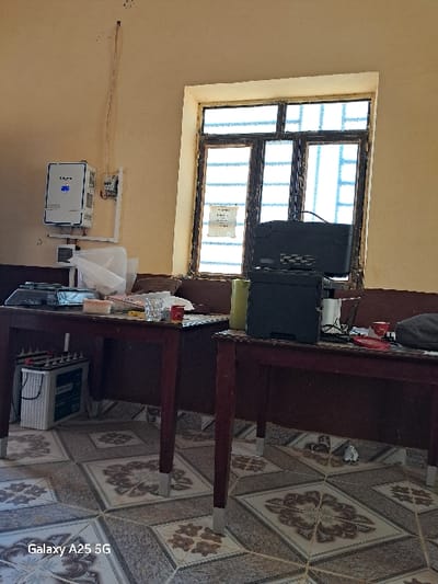 The image shows a room with a large window, two wooden desks, and a white wall. There is a black computer on the left desk, a battery on the floor next to it, and a solar panel on the wall. The window is barred and has a view of a cloudy sky. The floor is tiled with brown and white patterned tiles. The location is in the city of Karima, Sudan.