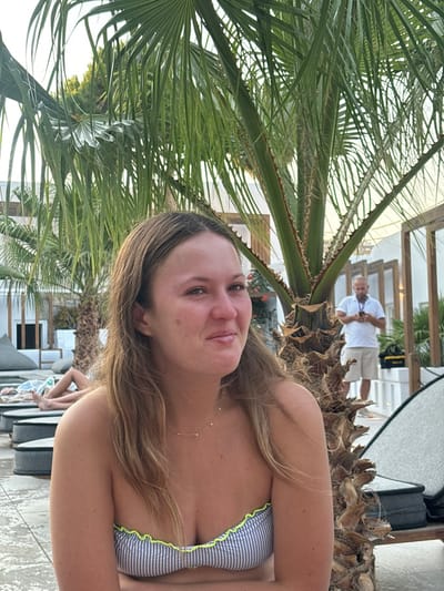 The image depicts an outdoor leisure area in Platys Gialos, Greece, illuminated by natural daylight.
A female individual, appearing in her 20s to early 30s, occupies the foreground. She has light brown hair and is wearing a strapless, horizontally striped blue and white bikini top with a neon yellow-green ruffled edge. A thin, gold-toned necklace with small triangular pendants is visible on her chest. She is positioned slightly angled from the camera, with her head turned slightly to the viewer's left, displaying a subtle smile.
Directly behind the female, a large palm tree with a robust, textured trunk and green fronds dominates the central background. To the left of the frame, partially visible bare legs of an individual are resting on a dark-cushioned outdoor lounge bed. To the right of the frame, positioned behind the central palm tree, a male individual is standing. He appears to be middle-aged, dressed in a white short-sleeved collared shirt and light-colored shorts. He is holding a smartphone with both hands and observing its screen. A yellow bag or object rests on the ground near his feet.
The environment consists of light-colored, possibly paved, ground. In the mid-ground and background, multiple white-washed buildings with wooden-framed windows and doorways are visible, consistent with a resort or hotel complex in a Mediterranean setting. Various dark-cushioned outdoor lounge furniture pieces are scattered throughout the scene.
