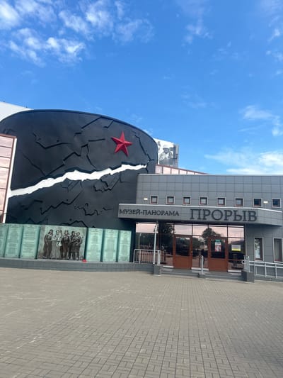 The image shows the Museum-Panorama "Prokhorovka" located in Shlisselburg, Russia. The museum features a large, black, curved facade decorated with a red star and a white jagged line, resembling a cracked surface. In front of the museum, a row of illuminated glass panels displays portraits of soldiers and lists of names. The entrance to the museum has double glass doors with signs indicating "ВЫХОД" (Exit) and "ВХОД" (Entrance). The surrounding area is paved with grey interlocking bricks. The sky is blue with scattered white clouds.