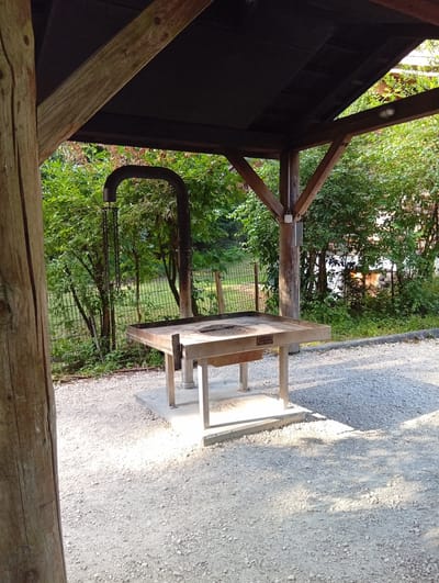 The image shows an outdoor grill station under a wooden shelter in Meiniswil, Switzerland. The grill is made of stainless steel and is situated on a concrete platform. It has a wide, flat cooking surface with a raised edge. A metal chimney extends upwards from the grill, curving over to the side. Chains hang from the chimney, presumably for adjusting the grill height or hanging cooking utensils. The shelter has a dark roof and wooden beams supporting it. The ground around the grill is covered with light-colored gravel. In the background, there is a metal fence, lush green trees, and foliage, suggesting a natural or park-like setting. The lighting indicates it is daytime, with shadows cast by the shelter and trees. There are no people visible in the image.