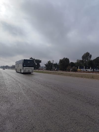 The image captures an exterior view of a wet, multi-lane asphalt road under a heavily overcast sky, located in Mankat al-Hatab, Syria. A white coach-style bus, displaying a Syrian license plate (٦١٥٦٨٧ / ١٤), occupies the right lane and is moving away from the camera. In the distant left lane, another vehicle is faintly visible, traveling towards the camera. The road surface exhibits clear signs of dampness or recent rain. To the right of the roadway, an unpaved median strip with sparse, dry vegetation separates the asphalt from a line of mature trees. Behind these trees, a large blue structure or sign with white lettering is partially visible, alongside multiple parked vehicles including several white vans, a yellow car, and other standard sedans. Utility poles with overhead lines are discernible amongst the tree line. The far horizon to the left reveals the indistinct outline of hills or mountains beneath the cloudy sky. No human figures are present in the frame.