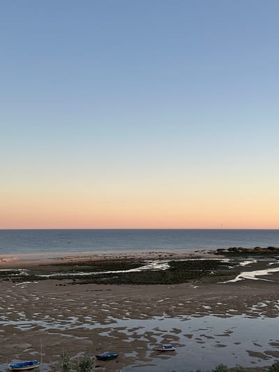 The image depicts a coastal scene at low tide. A wide expanse of wet sand is visible, reflecting the sky and creating mottled patterns. Two small boats are beached on the sand. A strip of vegetation runs along the edge of the sand. In the distance, the sea meets the sky, with a gradient of colors from blue to orange. The location is likely in Vila Real de Santo António, Portugal, as suggested by the prompt, given the coastal geography and tidal range.