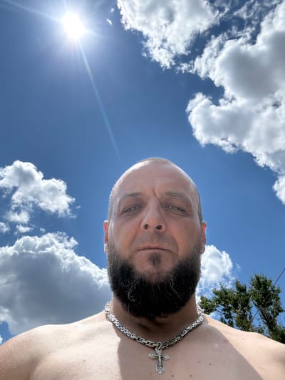 A low-angle, close-up, outdoor selfie shows a man with a bald head and a full beard, wearing a thick, silver chain with a cross pendant. He is looking directly at the camera. The background consists of a bright blue sky with scattered white clouds and the sun, which is casting lens flares. A portion of a green tree is visible on the right side of the frame. The man's shoulders and upper chest are visible, and his skin appears to have some texture. There is no indication of the city of Zhirnov, Russia in the image.