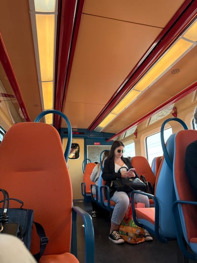 The interior of a passenger train car is depicted, operating in the city of Castanheira do Ribatejo e Cachoeiras, Portugal. A young adult female, with long dark hair, wearing sunglasses, a white top, a black blazer, light blue jeans, and black Converse-style sneakers, is seated on the right side of the aisle in an orange fabric seat with a blue frame. She holds a pink smartphone in both hands, actively viewing its screen, and simultaneously holds a white cup. A black shoulder bag is positioned on her lap, and a multi-colored plastic shopping bag rests on the dark floor between her feet. The train car features a light orange ceiling with multiple elongated, illuminated light fixtures bordered by dark red trim. Seating consists of orange fabric cushions with blue frames and armrests. Large windows are visible on the right, displaying an indistinct exterior. A blue door frame with a small window and a red digital display are visible on the left side of the car. Other passengers are partially visible in the background and foreground, seated.