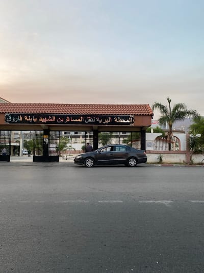 In Blida, Algeria, a dark sedan is parked in front of a bus station entrance. The bus station's facade features a tiled, orange roof and a black sign with red Arabic lettering. The sign appears to read "المحطة البرية لنقل المسافرين الشهيد بابلة فاروق" which translates to "The Land Station for the Transport of Passengers, the Martyr Babla Farouk." The entrance is secured by black metal gates. A person wearing a dark jacket is standing near the car, and other individuals are visible through the gates inside the station. A palm tree and other greenery are present, with a mountainous landscape in the background. The sky above is a soft gradient of pale blue and pink, indicating either dawn or dusk. The street in the foreground shows a dashed white line, typical of a roadway.
