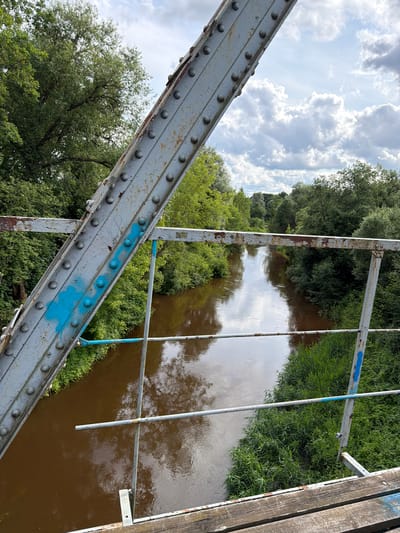 The image depicts a view from a weathered metal bridge looking down at a brown river flowing through a verdant landscape. The bridge's structure, composed of large, riveted metal beams, shows signs of age with peeling paint and rust. Blue spray paint is visible on one of the bridge's main supports. The riverbanks are densely covered with lush green trees and vegetation. The sky above is partly cloudy, with a mix of white, fluffy clouds and patches of blue. The water reflects the cloudy sky and the surrounding greenery. The scene appears to be an outdoor, natural setting, likely in a rural or semi-rural area. This scene is located in the vicinity of Tēraudi, Latvia, a region characterized by its natural beauty and waterways. There are no people visible in the image.