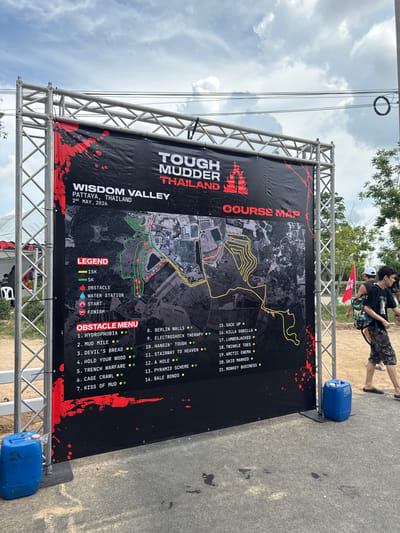 Here is a detailed description of the image:
**Scene:**
The image shows a banner for a "Tough Mudder Thailand" event. The event is to be held at Wisdom Valley in Pattaya, Thailand, on May 2nd, 2026. The banner is held up by a metal frame and secured with blue jugs at the bottom.
**Banner Details:**
The banner displays the event's name, location, and date. A course map is prominently featured on the banner. The map outlines both a 15K and 5K course, marked by yellow and green lines respectively. The legend details that red markers indicate obstacles, blue markers denote water stations, a red circle indicates the start, and the red intertwined rings indicate the finish line. The course map is overlaid on what appears to be a satellite image of the Wisdom Valley area. An "Obstacle Menu" lists the various obstacles along the course, labeled numerically.
**Setting/Environment:**
The banner is set up on a paved road. The background features trees and a cloudy sky.
**People:**
Part of two people can be seen in the upper-right corner of the image. One is wearing a black shirt and camouflage pants and carrying a blue shoulder bag, while the other one is wearing a white hat and black backpack.
**Location:**
The image is most likely taken in the vicinity of Wisdom Valley in Pattaya, within the city of บ้านสำนักตะแบก, Thailand.
**Additional details:**
The background is overcast, suggesting cloudy weather during the photograph.