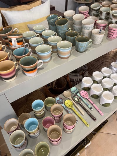 The image shows a retail display of ceramic mugs and spoons on a light gray shelf. The mugs are arranged in rows and stacks, with a variety of colors and patterns. Some mugs have a speckled glaze, while others have horizontal stripes. The spoons are laid out in a row on the shelf, and they also feature various colors and patterns. In the background, there are pillows and woven baskets, suggesting a home decor or gift shop setting. The location is identified as Pollença, Spain, implying this is a scene from a shop in that town. There are no people visible in the image, and no specific actions are taking place. The focus is entirely on the product display.