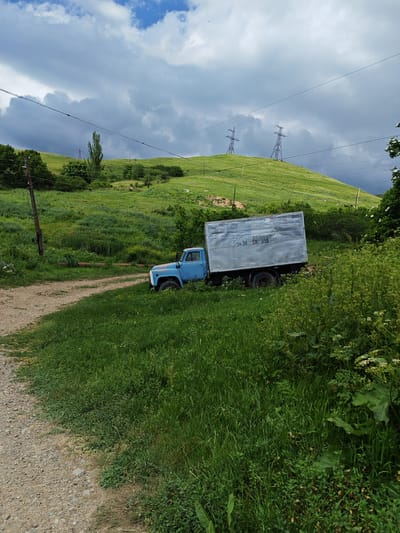 The image depicts a light blue, aged truck with a silver cargo box parked on the right side of a dirt road. The truck is partially overgrown with tall green grass and vegetation. The road curves to the left and leads uphill towards a grassy hillside. In the background, two tall electrical pylons stand on the crest of the hill. The sky is partly cloudy with a mix of gray and white clouds. The environment appears to be rural, with rolling green hills and scattered trees. The text "AM 34 GA 915" is visible on the side of the truck's cargo box. The scene is likely located in or near the city of Bazum, Armenia, given the context. There are no people or animals visible in the image.