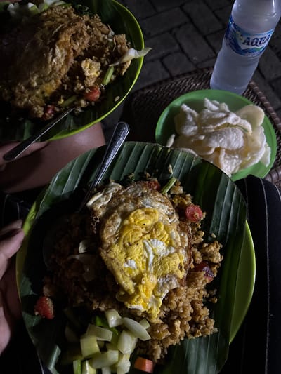 This image captures a casual night meal, likely outdoors or in a dimly lit setting, consistent with a street food stall or warung in Indonesia. Two servings of Nasi Goreng (fried rice) are presented on green plastic plates, each lined with a fresh banana leaf, a traditional method of serving. Both plates of fried rice are topped with a fried egg or omelet and garnished with vegetables like chopped tomatoes and possibly some greens or cabbage. One plate also features a side of pickled cucumber and carrot salad (acar).

Adjacent to the main dishes, a smaller green plate holds a generous pile of white, airy crackers (kerupuk). A plastic bottle of "AQUA" brand mineral water, showing "600ml" and other small text, is visible in the upper left corner. Metal spoons rest on the banana leaves next to the fried rice.

The scene is dimly lit, suggesting it is nighttime, with strong shadows cast by a direct light source. The background shows a dark, possibly paved ground or floor with a brick pattern, and a woven surface, likely a table or mat. Parts of a person's hands and legs, clad in dark clothing, are visible, indicating diners are present and about to enjoy the meal. The overall atmosphere is unpretentious and focused on the food.