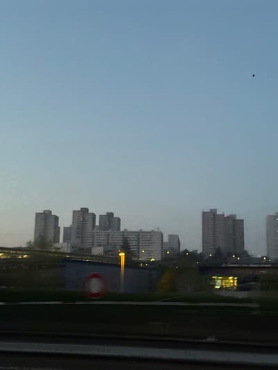The image depicts an urban landscape in Noisy-le-Sec, France, at dusk or dawn. The sky is a gradient of light to dark blue. A small, dark, winged object, possibly a bird, is visible in the upper right quadrant of the sky. The horizon features a cluster of multi-story, utilitarian-style buildings, appearing as silhouettes against the dim light, with some internal illumination visible in select windows. In the immediate foreground, a dark, horizontal band suggests railway tracks or a road. Above this, a dark, vegetated embankment or barrier is visible. A blue wall or structure stands in the mid-left, bearing a red and white circular object, resembling a lifebuoy or warning sign. An active, yellow-orange street lamp stands illuminated on the left, casting light. Further to the right, another light source and interior illumination from a building are discernible. No people or direct interactions are visible.