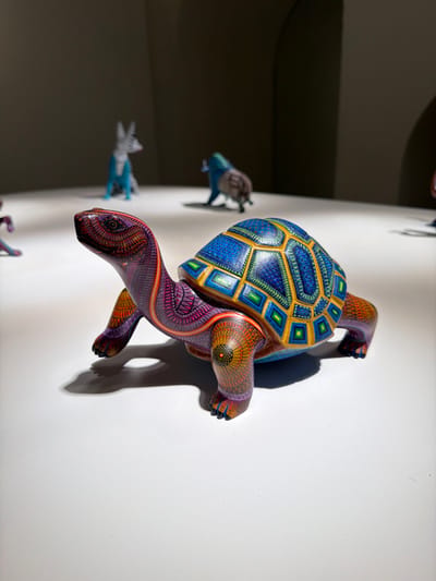 A vibrant, hand-carved turtle sculpture clings to a white wall. Its shell boasts intricate blue, yellow, and green geometric patterns, while the head and limbs display a rich blend of purple and orange hues. Its shadow casts a dark silhouette on the wall behind it. Other animal sculptures are visible in the background on the same wall, adding depth to the image.
