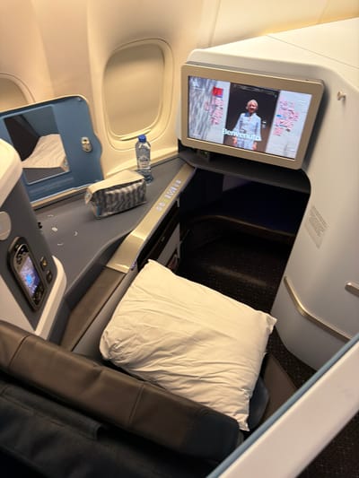 The image depicts the interior of a business class airplane seat. The seat is reclined and features a dark leather armrest. A white pillow is placed on the seat. To the left of the seat, there is a dark grey surface with a built-in screen displaying a person, and what appears to be a control panel. A bottle of water and a toiletries bag are also present on this surface. The background shows a window and the wall of the airplane cabin. The scene is well-lit, suggesting it is inside the cabin during daytime or with artificial lighting. This appears to be a static, posed shot rather than an action shot. There are no people visible in the image. The location within Schiphol, Netherlands is not discernible from the image itself.