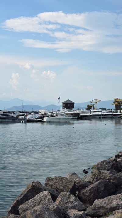 The image depicts a serene scene at a marina in Peschiera del Garda, Italy. The marina is lined with several white boats of varying sizes, docked neatly along the waterfront. In the foreground, to the right, large, textured rocks form a natural embankment, with a dark bird perched on one of them. The water of the marina is calm, reflecting the pale blue sky dotted with wispy white clouds.

In the background, a small, dark-colored building with a pointed roof stands on a jetty, with a flag flying beside it. Beyond the marina, a hazy mountain range forms a picturesque backdrop, suggesting a clear day with good visibility. A yellow crane is visible further down the marina, hinting at ongoing port activities or construction.

The overall atmosphere is tranquil and peaceful, evoking a sense of calm summer day. The lighting suggests it is daytime, likely late morning or early afternoon, given the brightness and the presence of shadows. The text "POLIZIA PROVINCIALE" is visible on the side of one of the boats, indicating it is a police vessel. The scene is typical of a lakeside or coastal town in Italy, consistent with the provided location context of Peschiera del Garda.