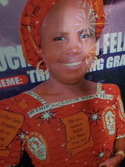 This media file features a portrait of a woman adorned in a vibrant red and orange patterned fabric, likely a traditional Nigerian attire. The fabric is rich with intricate designs, including starbursts and what appear to be religious emblems. One emblem shows "Church Of Christ In Nations COCIN". Another section of the fabric displays a quote in a scroll-like design: "The wise woman Builds her House™ Prov. 1:1".

The woman's face is partially visible, with her eyes looking towards the viewer. She has a warm, gentle expression. The background of the image is dark, with some blurry text and colorful, abstract graphics that suggest a celebratory or commemorative event, possibly a poster or banner. Text visible on a dark blue background reads "NG GRA" and "EME: TH". There are also words like "COCIN WOMEN FELLO", "SHIP", and "La Plus Sage Femmes".

The overall impression is one of pride and cultural significance, possibly related to a church event or a celebration honoring wise women within the community. The setting appears to be indoors, given the controlled lighting and the nature of the backdrop. The woman is wearing gold bangles on her wrist. The image is slightly tilted, adding a dynamic feel.