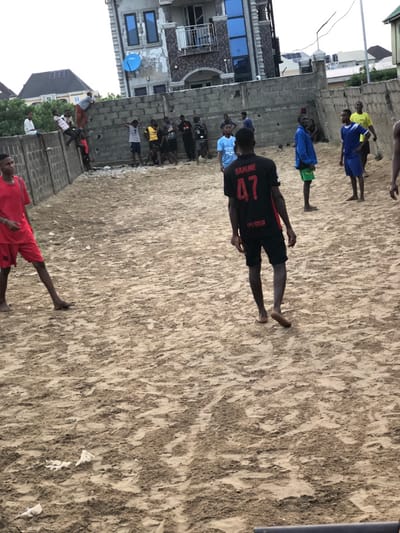 A group of people are playing on a sandy area near a building. Many are barefoot. One person wears a black jersey with "SAMME 47" on the back. Another individual wears a red outfit and is in the lower portion of the scene. Others are wearing blue and yellow athletic wear. The sand appears uneven and textured. A building stands to the side with windows and a satellite dish.
