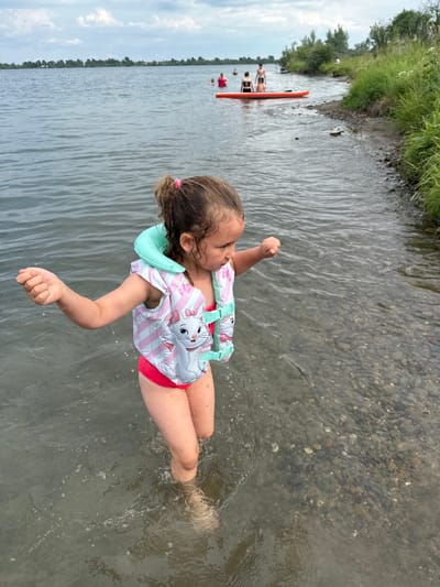 A young girl wearing a pink and white striped life vest with a cartoon cat on it, and red swim bottoms, is in shallow water. She has her arms raised and appears to be kicking her legs, creating ripples in the water. Her hair is light brown and tied with a pink scrunchie. The water is clear enough to see the pebbly bottom. In the background, a person is on an orange paddleboard, and other people are in the water. The scene takes place outdoors on a cloudy day, likely in the afternoon, at a lake or river with a grassy bank and trees in the distance. The overall mood is one of summer fun and recreation.