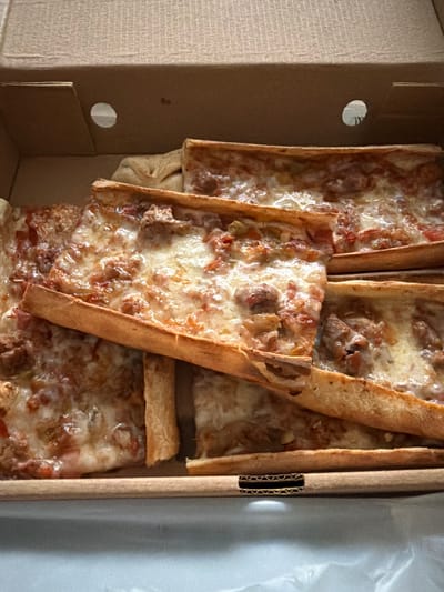 This is a top-down, close-up shot of a rectangular pizza in a cardboard box. The pizza is cut into several slices, and the toppings appear to be cheese, tomato sauce, and some kind of meat, possibly sausage. The pizza is layered, with some slices placed on top of others. The cardboard box is light brown and appears to be slightly worn. The background is out of focus. The image was taken in Zeytinburnu, Turkey.